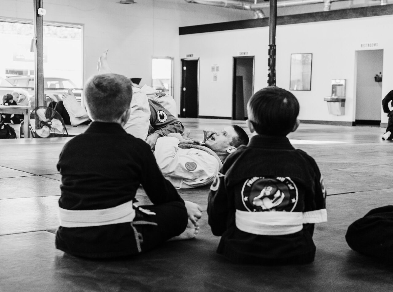 Two young boys in Jiu Jitsu gis focus on black belt professor Matt Vernon as he demonstrates a submission technique during kids BJJ class at Primate Jiu Jitsu in Tulsa OK