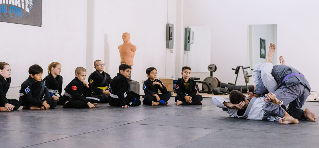 A group of disciplined and hardworking children sit quietly and focus as they watch black belt professor Matt Vernon and purple belt assistant kids coach Chris Stafford demonstrate submission grappling techniques in the gi kimono during a kids BJJ class at Primate Jiu Jitsu in Tulsa OK