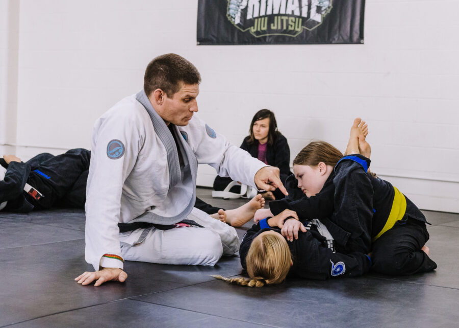 Black belt professor Matt Vernon teaches young girls in kids BJJ class at Primate Jiu Jitsu how to properly secure and finish the triangle choke strangle submission