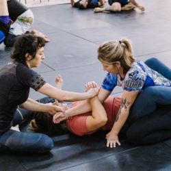 Black belt coach Kendall Vernon shows women at Primate Jiu Jitsu how to properly secure the arm for an arm bar attack from closed guard during an adult nogi submission grappling class at Primate Jiu Jitsu while wearing Gaidama