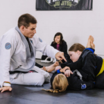 Black Belt Professor Matt Vernon coaches two girls during kids BJJ class at Primate Jiu Jitsu in Tulsa OK offering details for finishing the triangle submission